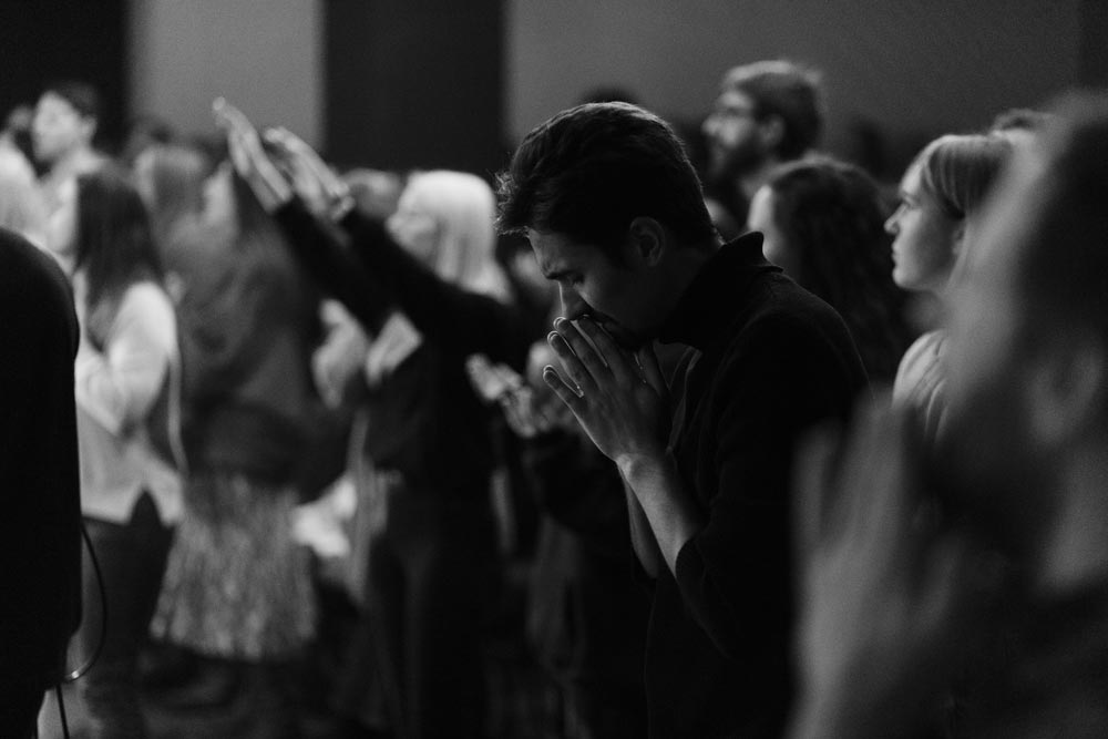 Group of people in a worship service with eyes closed and hands lifted, focusing in prayer and reverence before God