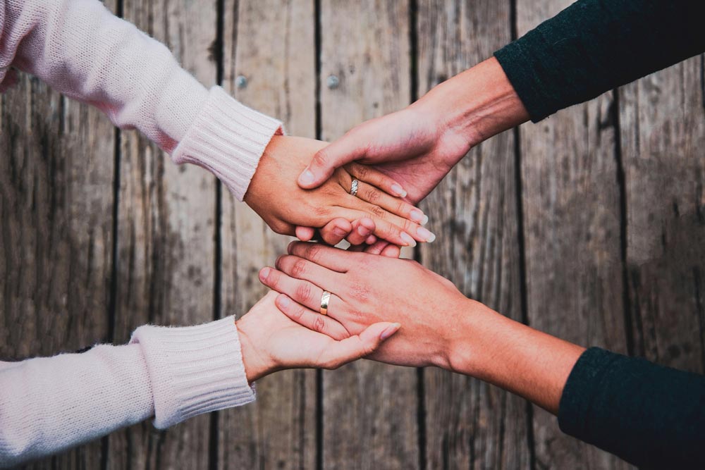 Close-up of four hands clasped in prayer and unity over a rustic wooden surface, representing agreement and support in faith